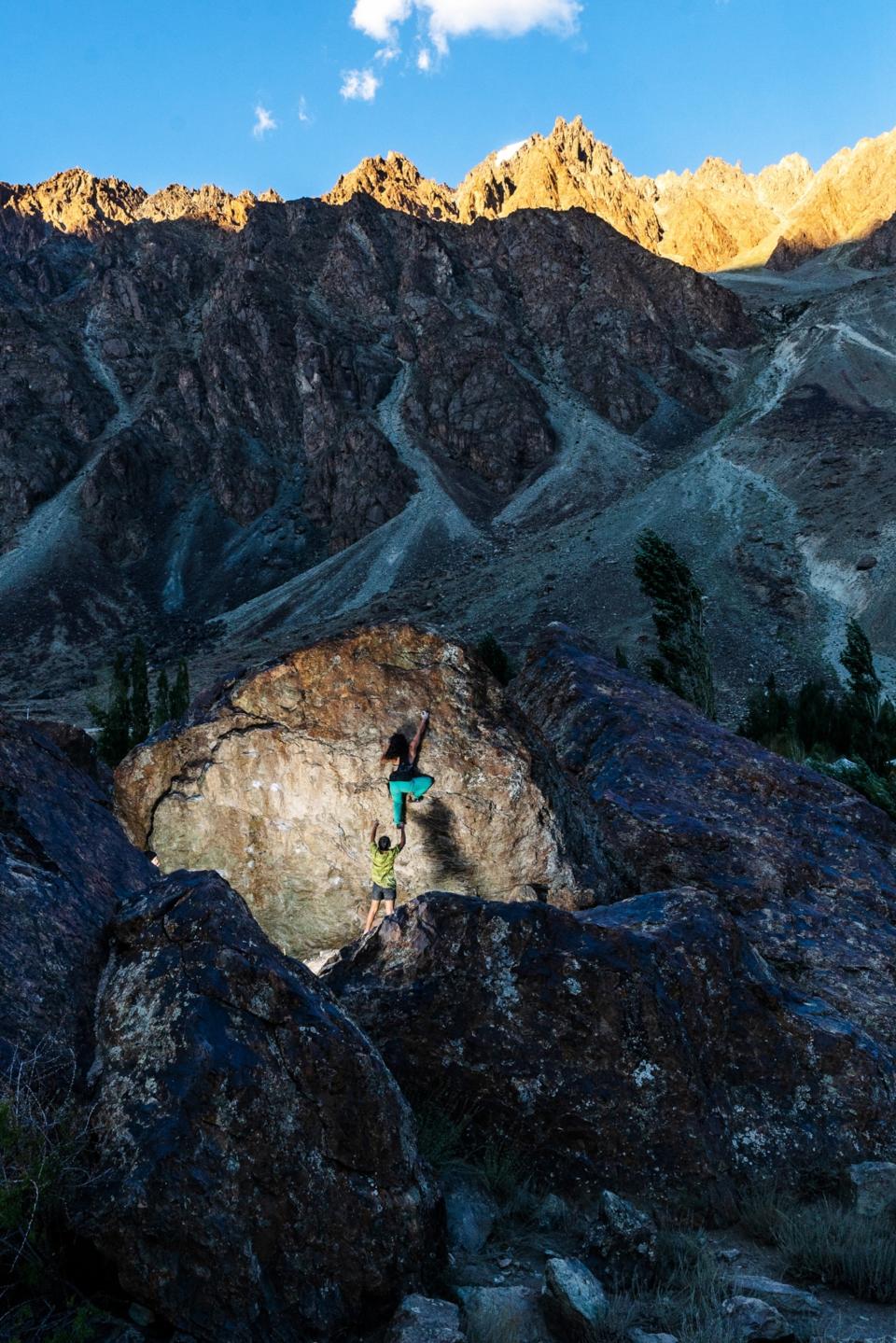 A rocky valley with mountain peaks kissed by sunlight in the background. Near the foreground, a camera flash shines on a woman climbing the sheer face of a boulder as another woman stands by as a spotter.