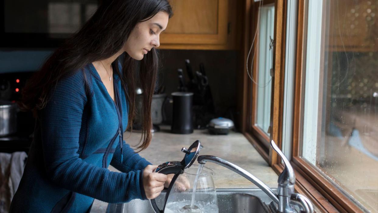 Young woman with long dark hair fills up a clear kettle with water at a kitchen sink
