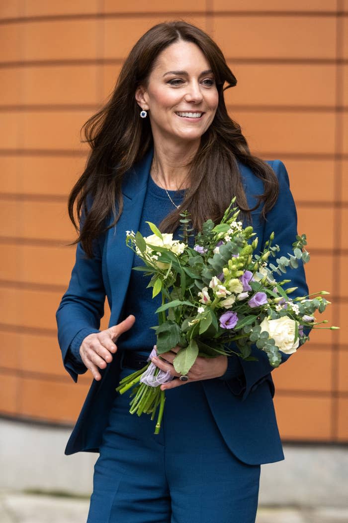 Kate Middleton smiling, wearing a blue blazer, holding a bouquet. She's outside, near a brick wall