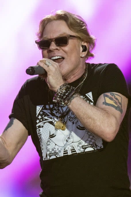 A person in a black graphic t-shirt and sunglasses sings passionately on stage, holding a microphone with a colorful background