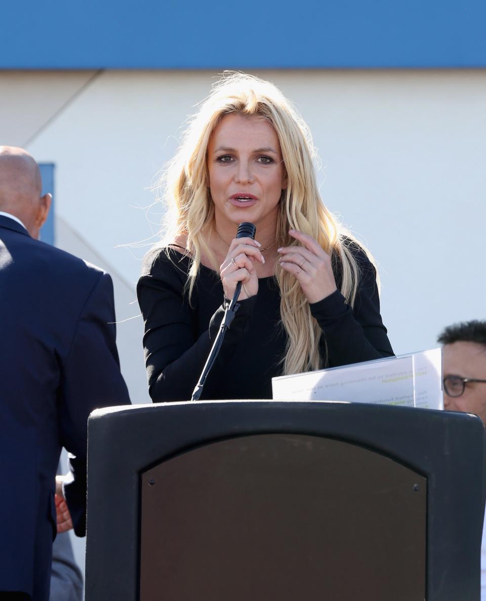 Britney Spears speaking into a microphone at an outdoor event, standing behind a lectern