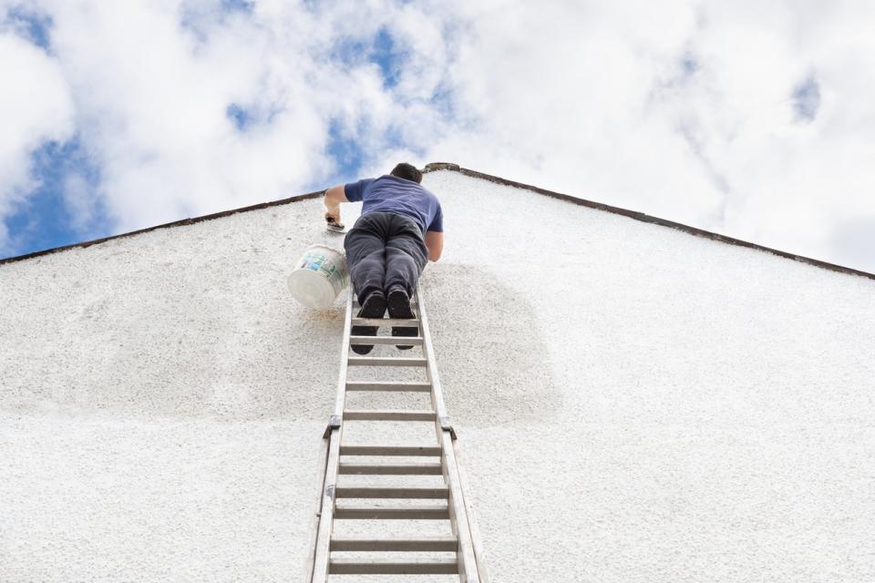 High quality photo of a man up high on a ladder painting a white pebbledash wall on the gable end of a house.