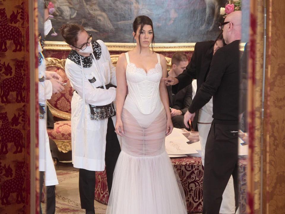 kourtney kardsahian standing in front of a mirror being fitted for a wedding dress. there are people around her tailoring in real time, while she stands stationary. the dress features a lacy bust, a unitard, and a sheer skirt