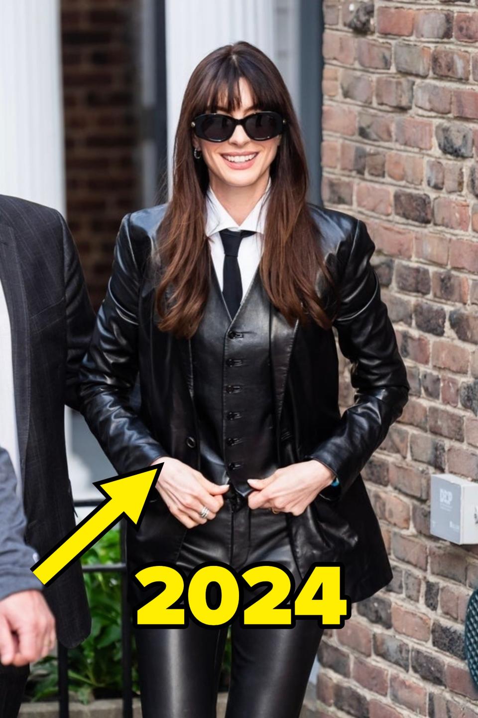 Anne Hathaway is outdoors, smiling, wearing a black leather jacket, black vest and pants, white shirt, black tie, and dark sunglasses