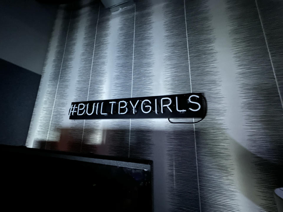 <p>A wide-angle photo from the iPhone 14 showing an unlit neon sign making out the words "Built By Girl" on a striped wallpaper.</p>
