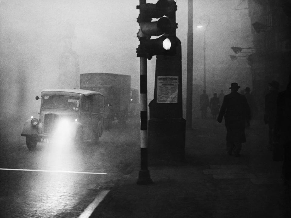 Traffic moves slowly, with lights aglow, as smog descends over British capital during daytime hours.