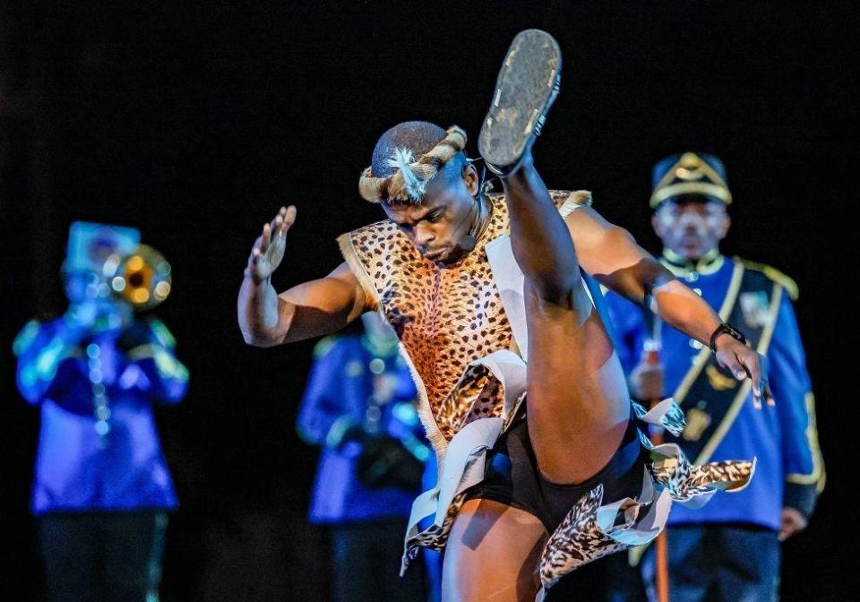 Members of the South African Force Band perform during the opening of the International Military Music Festival 'Spasskaya Tower' on the Red Square in Moscow. In the foreground of the picture is a male dancer energetically kicking his leg up high in the air, wearing a leopard-skin outfit and headdress. In the background are members of the South African Force Band. One is playing a trumpet. They are wearing blue jackets with gold trims.