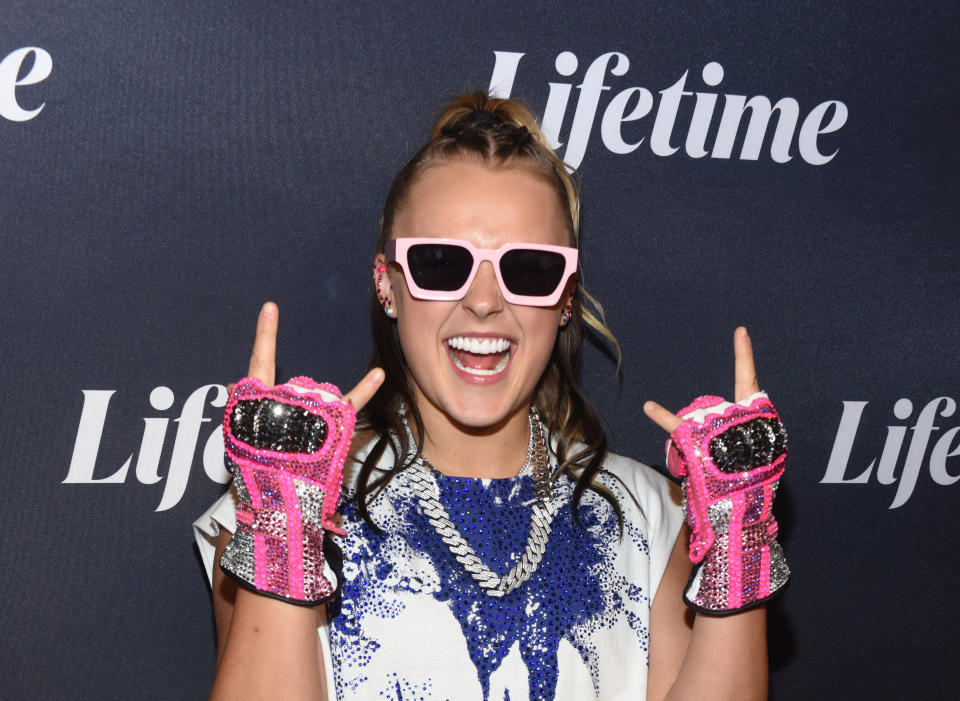 JoJo Siwa is seen smiling enthusiastically as she poses in front of a Lifetime backdrop wearing pink sunglasses, sequined gloves and a statement necklace.