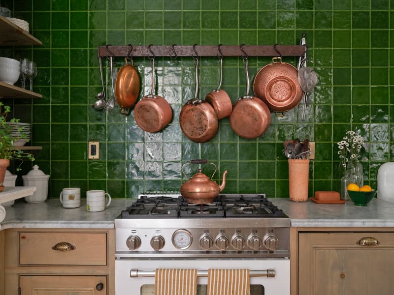 Copper pots hung above gas range in kitchen with green tiles.