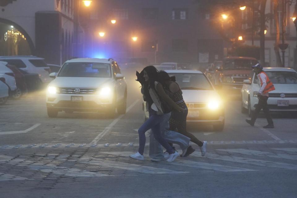 People cross a street where two vehicles with headlights on are stopped before a crosswalk