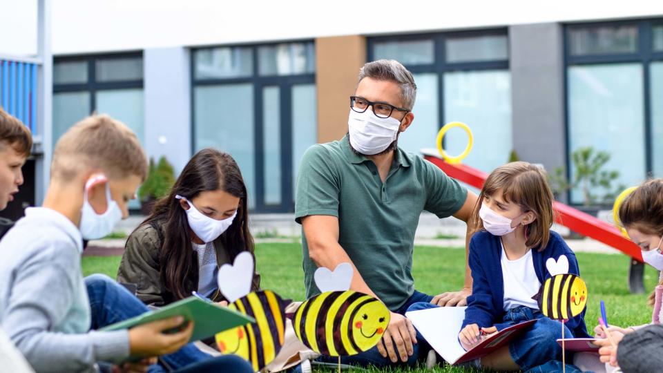 Group of cheerful children with teacher and face masks learning outdoors at school after covid-19 quarantine and lockdown.