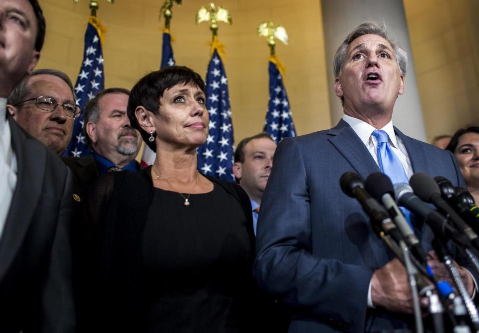 judy mccarthy stands next to kevin mccarthy who is speaking into several microphones, several people surround them, she wears a black outfit and jewelry, he wears a blue suit and tie