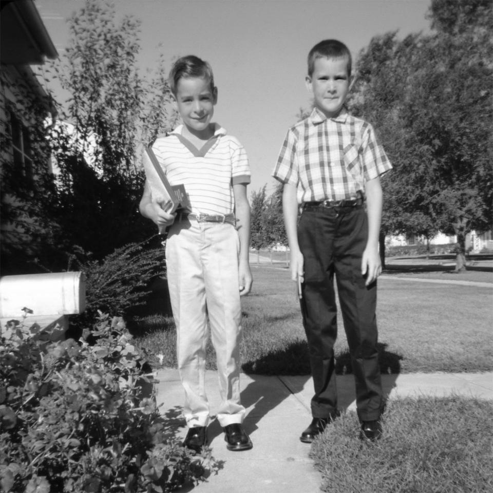 Two young boys, one with a striped shirt and book, and the other in a plaid shirt, stand on a sidewalk in a residential neighborhood