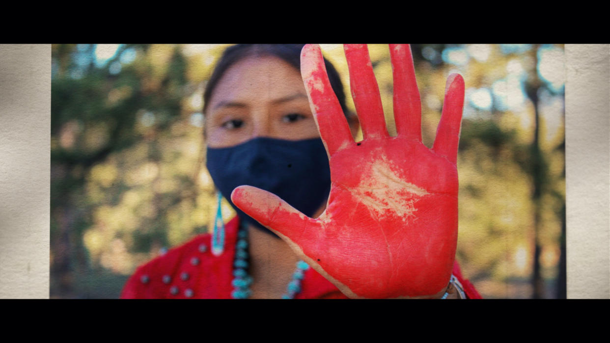 A young Native American woman holds out her open palm, painted red, toward the camera in the foreground.