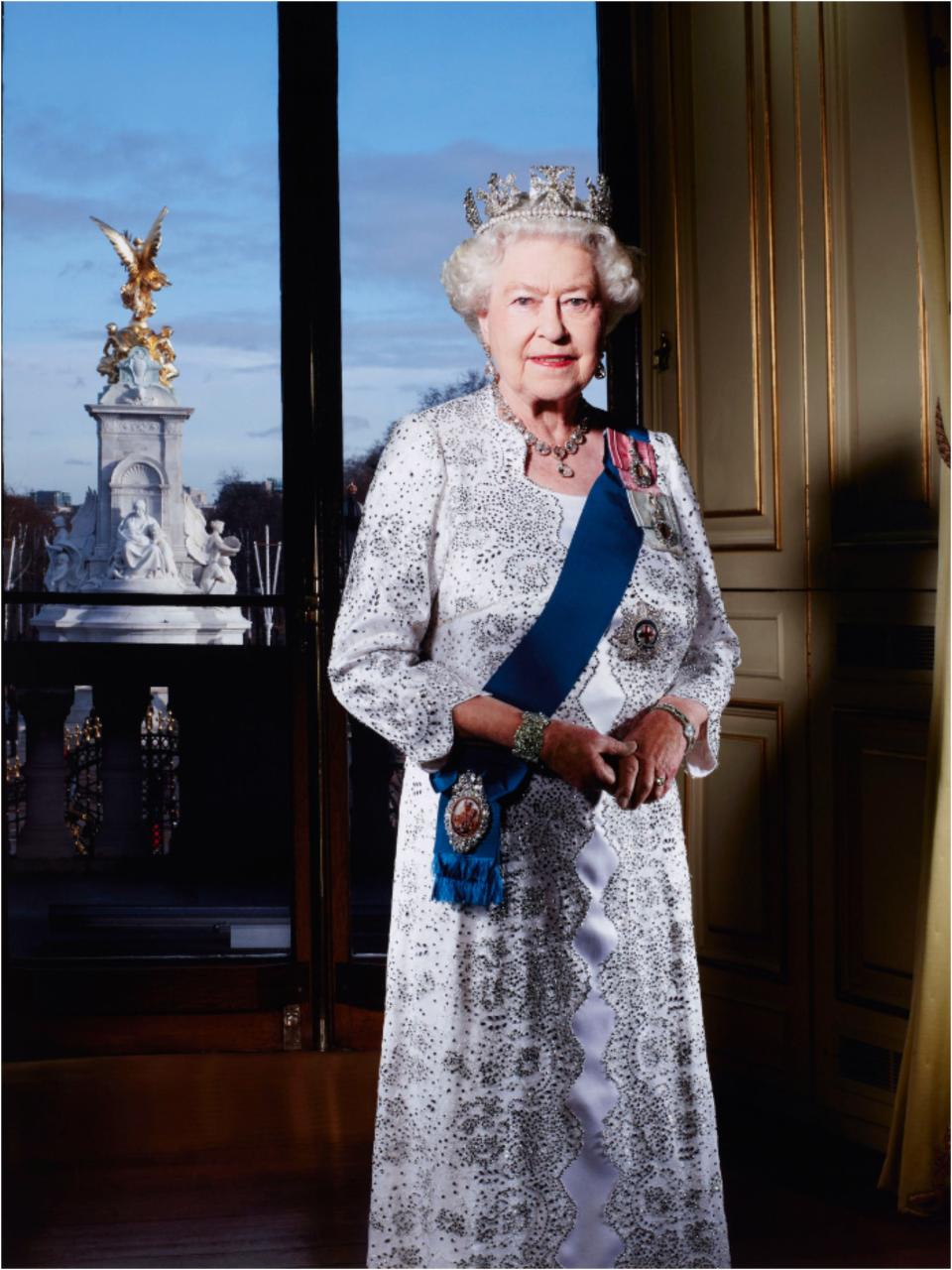 The Queen smiling in a white and silver brocade gown with a blue sash, medals, silver jewelry, and a delicate crown.