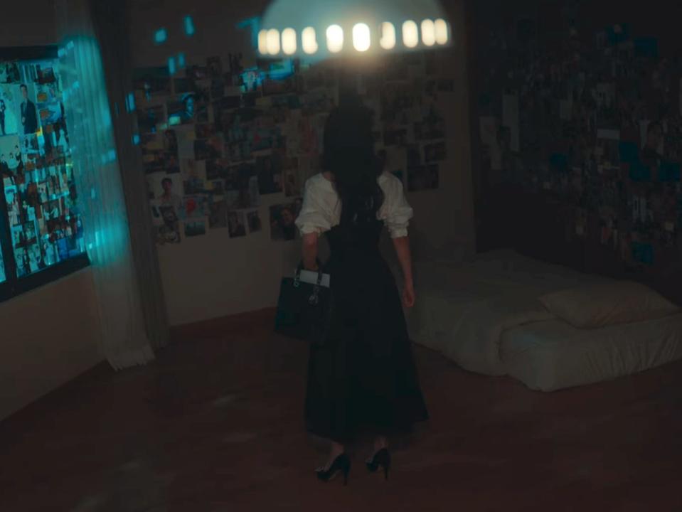 yeon-jin in the glory standing inside dong-eun's dark apartment. the walls are plastered with photos of yeon-jin and her friends, and there's a mattress with minimal bedding present in the corner of the room. yeon-jin is wearing her green heels