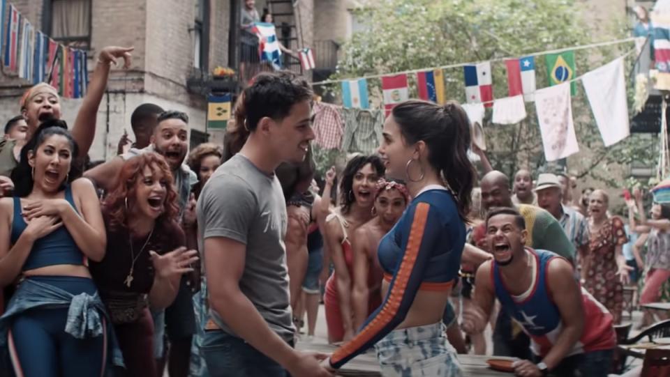 A man and a woman hold hands outside while a large group surrounds them and cheers them on in Warner Bros. In the Heights