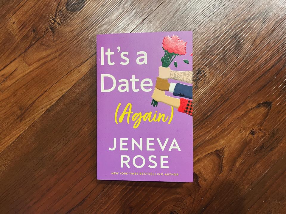 A book titled "It's a Date (Again)" by Geneva Rose lays on a wooden surface