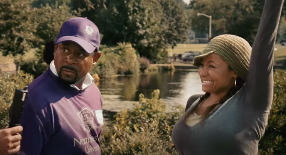 Two characters from a film, a man and a woman, stand outdoors with the man holding a fishing rod and the woman raising her arm in excitement