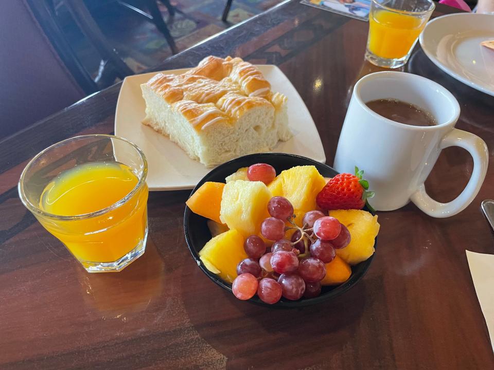 A glass cup of passion fruit, orange, and guava juice, a mug of coffee, a bowl of fruit, and plate of pineapple-coconut breakfast bread sit on the table.