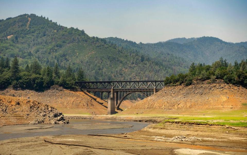 Forested mountains overlook a dry lakebed. A trestle bridge over mud and rock connects two hills.