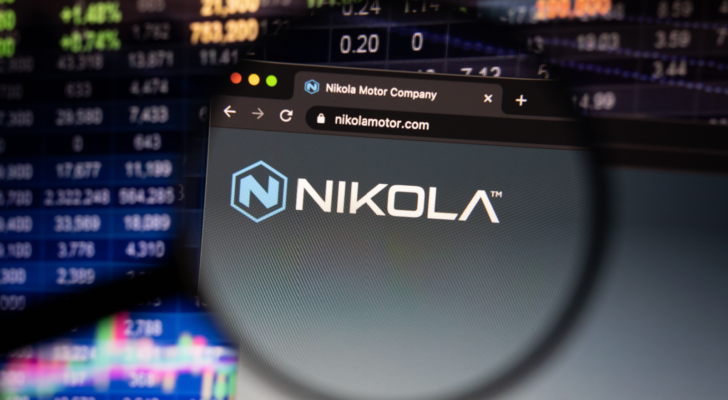 Nikola (NKLA) company logo on a website with blurry stock market developments in the background, seen on a computer screen through a magnifying glass.