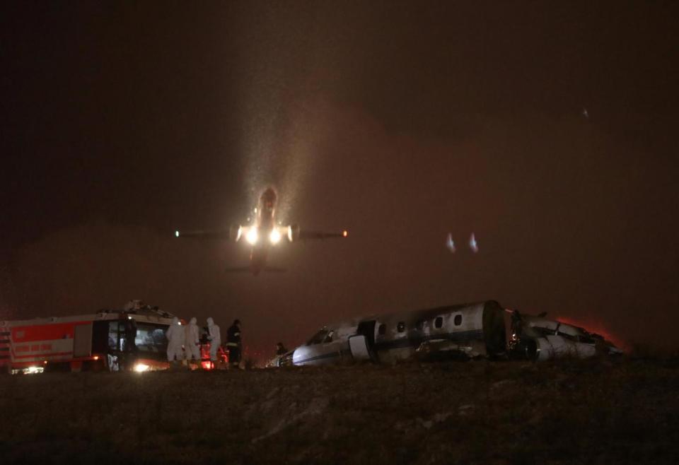 A passenger aircraft flies over the runway as firefigters stand next to the wreckage of a private jet (REUTERS)