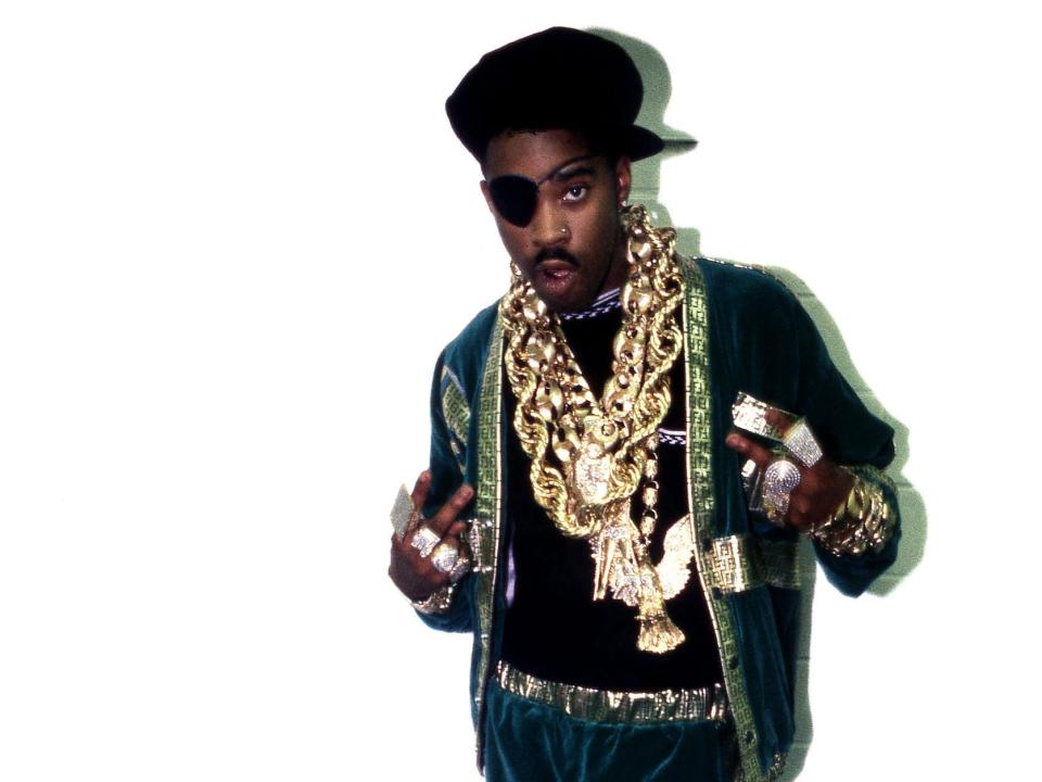 A photo of slick rick posing against a white wall and wearing many chains.