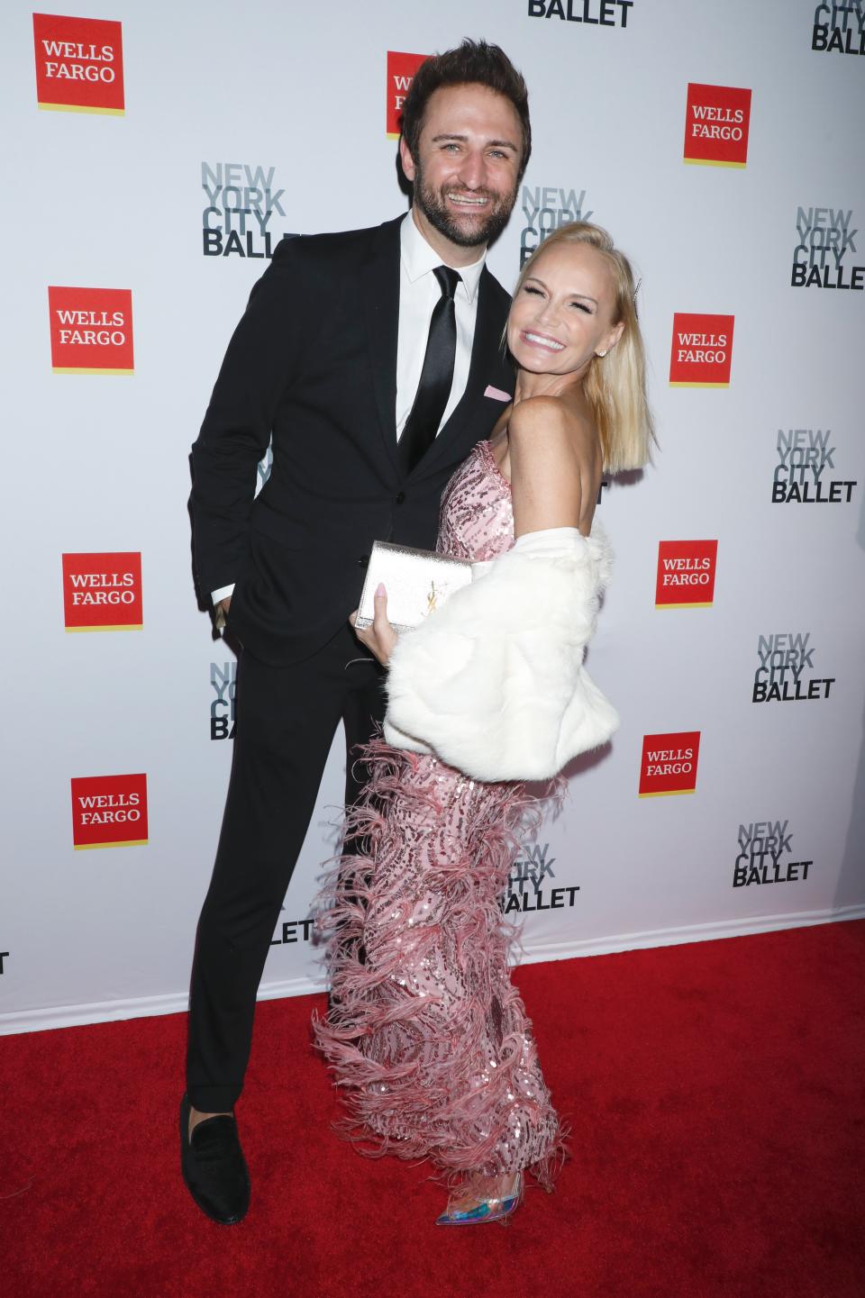 Josh Bryant smiles in a black suit and hugs fiancee Kristin Chenoweth, who wears a pink dress and white wrap