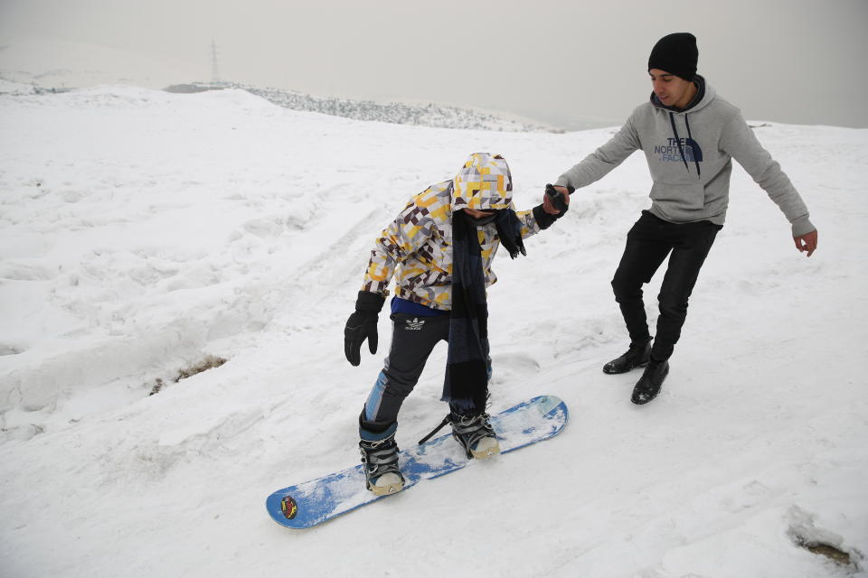 In this Friday, Jan. 24, 2020 photo, a member of the minority Hazara ethnic group, tries snowboarding with a trainer on the snow-covered hillside known as Kohe Koregh, on the outskirts of Kabul, Afghanistan. A handful of Afghanistan Snowboarding Federation members meet each weekend on a snowy slope above the city, encouraging others to give snowboarding a try — one run at a time. (AP Photo/Rahmat Gul)