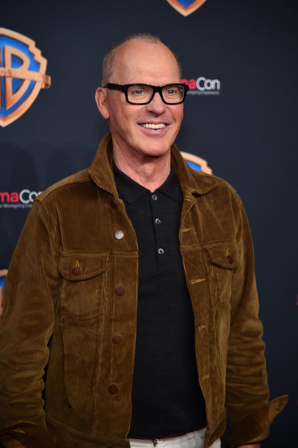 Michael Keaton smiling in a brown corduroy jacket and black shirt at Warner Bros. event at CinemaCon