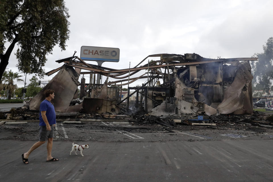 A man passes a bank burned during a protest over the death of George Floyd, Sunday, May 31, 2020, in La Mesa, Calif. A widely-seen video of a white police officer pushing the black man was taken after the officer stopped him for smoking on a trolley platform, authorities said Wednesday, providing additional details on what led to widespread damage during a protest days later. (AP Photo/Gregory Bull)