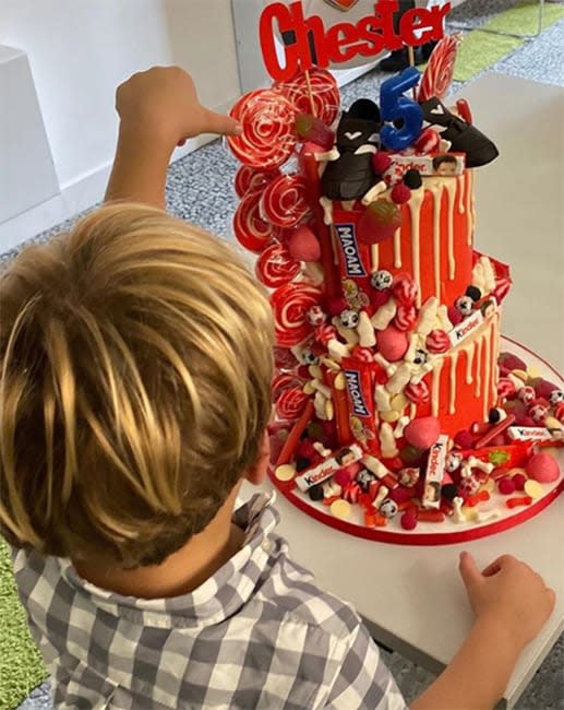 a photo showing a tall red and white stripy cake with a blond haired boy reaching for the top of it with his back turned to the camera