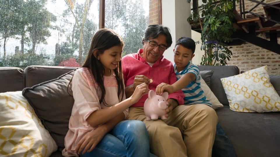 Grandfather teaching his grandkids to save money in piggy bank while sitting on couch at home all smiling.