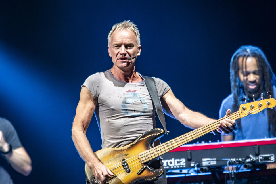 Un musicien en t-shirt gris joue de la guitare basse sur scène avec les membres du groupe en arrière-plan