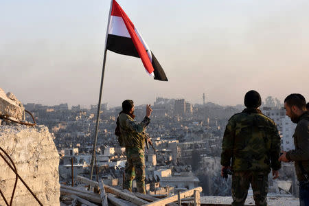 A Syrian government soldier gestures a v-sign under the Syrian national flag near a general view of eastern Aleppo after they took control of al-Sakhour neigbourhood in Aleppo, Syria in this handout picture provided by SANA on November 28, 2016. SANA/Handout via REUTERS
