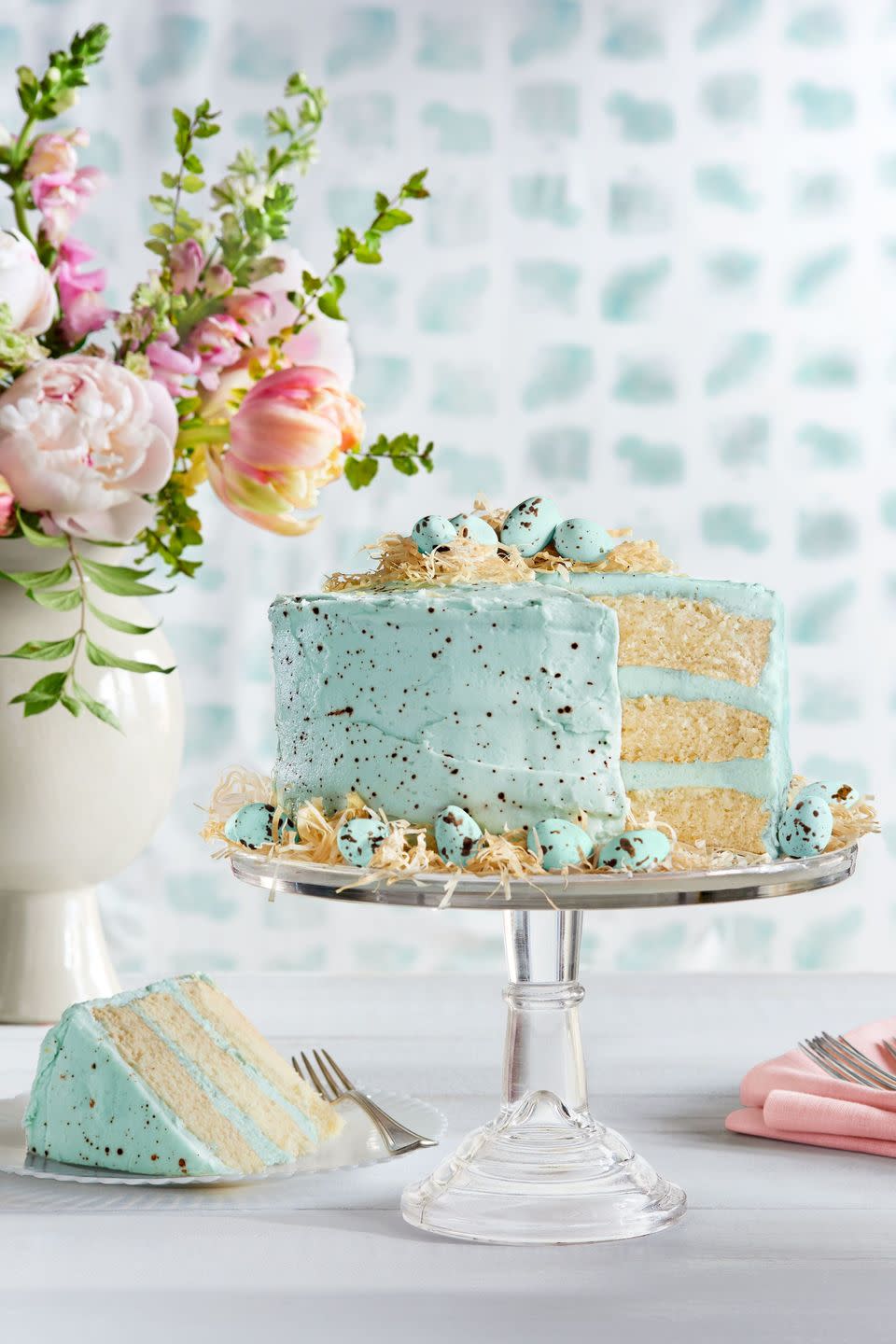 speckled malted coconut cake on a glass cake stand and garnished with speckled candy eggs and toasted coconut hay