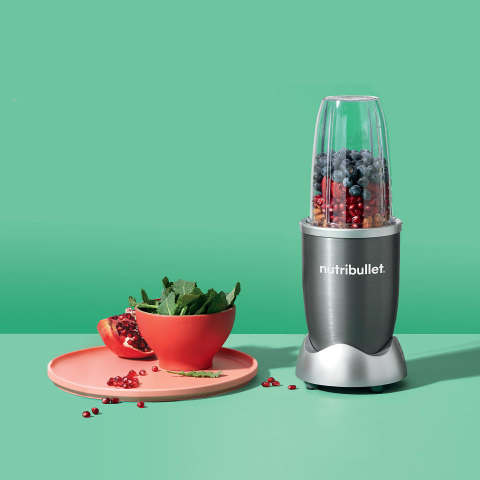 grey nutribullet with fruit inside next to a plate of vegetables and fruit on a green backdrop