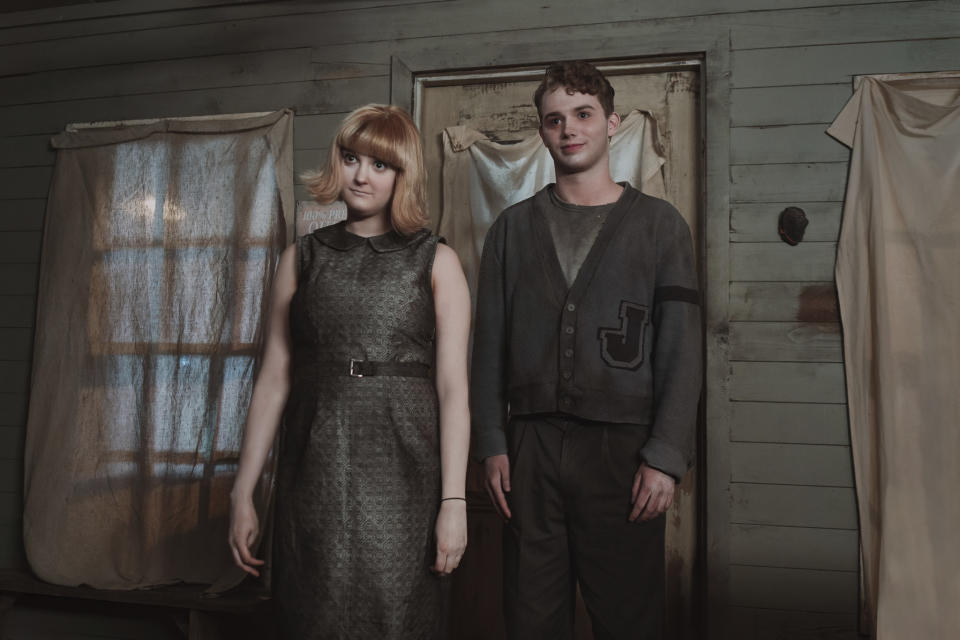 Two creepy pale figures in dirty, outdated clothes stand in front of a doorway