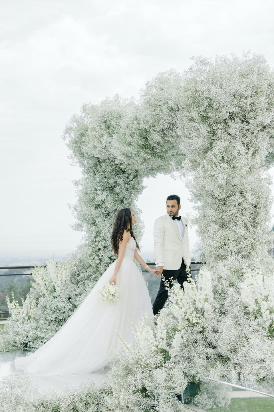 A bride and groom walk through a flower arch and smile at each other.