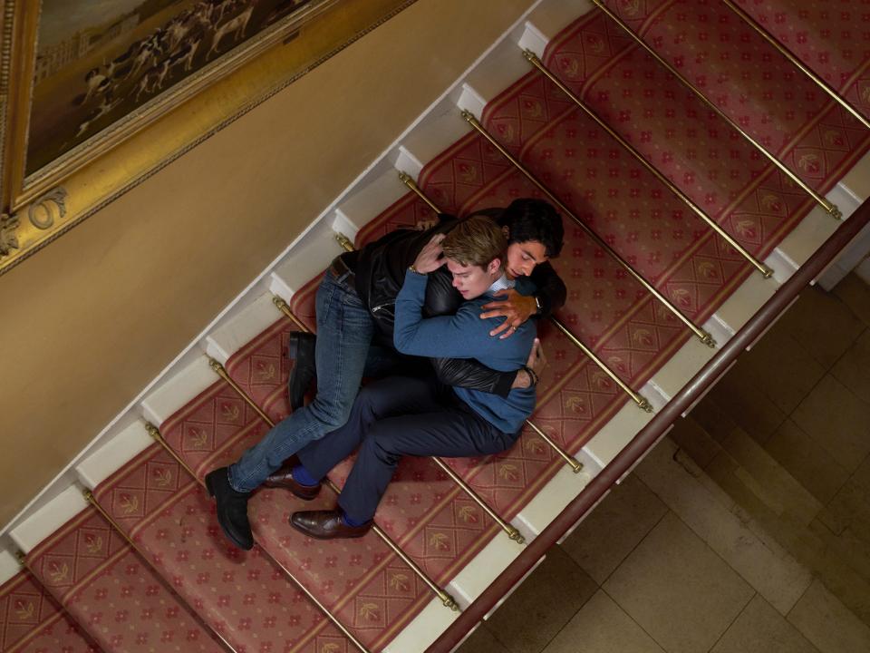 alex holds henry close to his body in red white and royal blue as they lay together on the stairs, clinging to each other