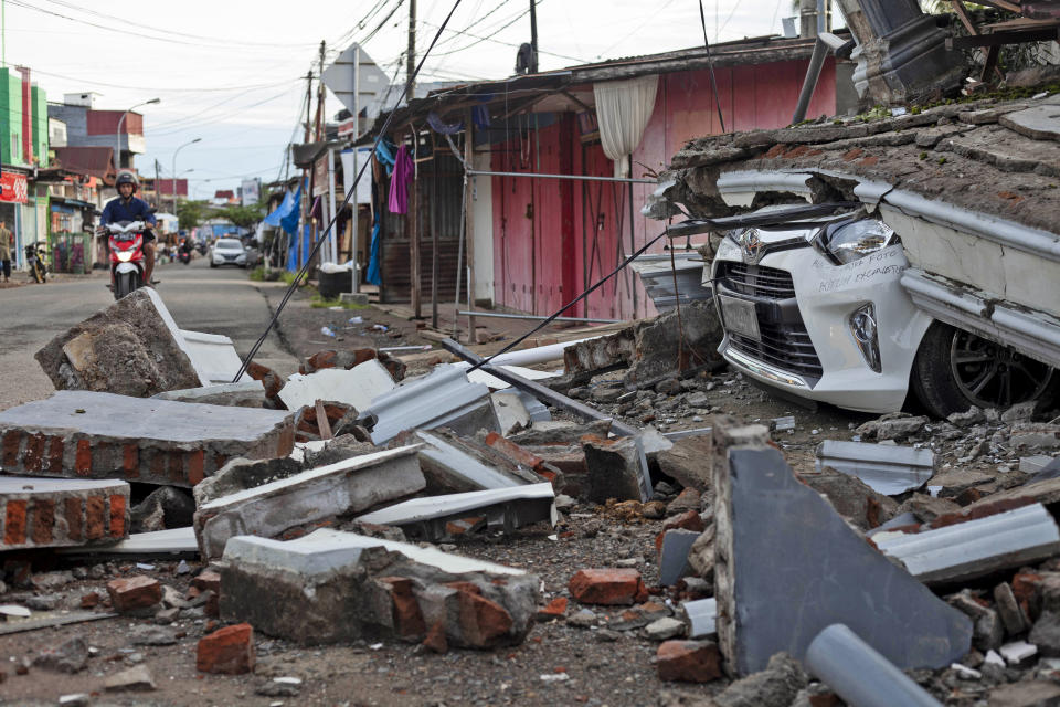 A motorist rides past the wreckage of a car buried under the rubble of a building collapsed in Friday's earthquake in Mamuju, West Sulawesi, Indonesia, Monday, Jan. 18, 2021. Aid was reaching the thousands of people left homeless and struggling after an earthquake that killed a number of people in the province where rescuers intensified their work Monday to find those buried in the rubble. (AP Photo/Yusuf Wahil)