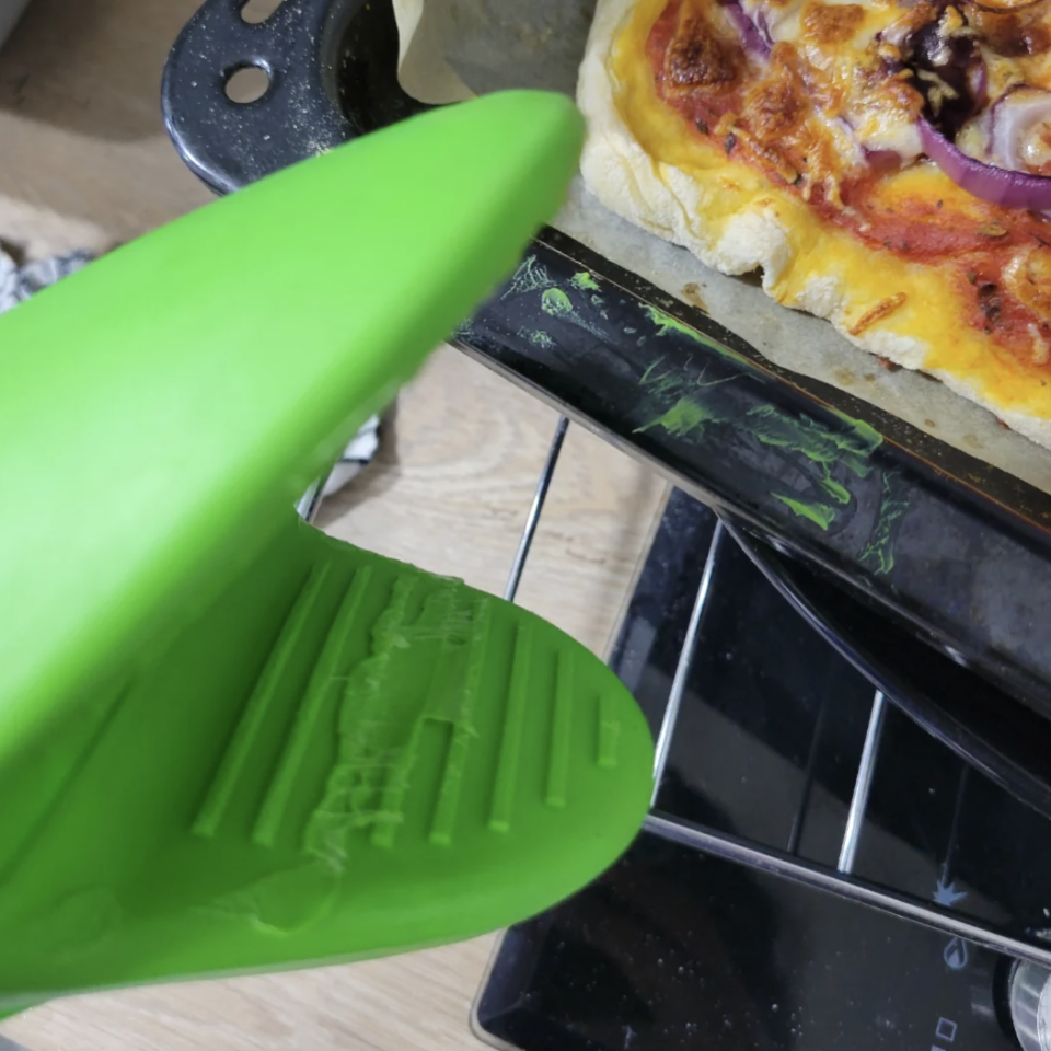 Hand in a green oven mitt with a melted section removing a pizza from an oven