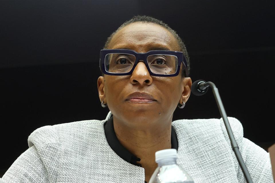 Claudine Gay speaks confidently into a microphone and is wearing a white suit and chunky black square glasses. 