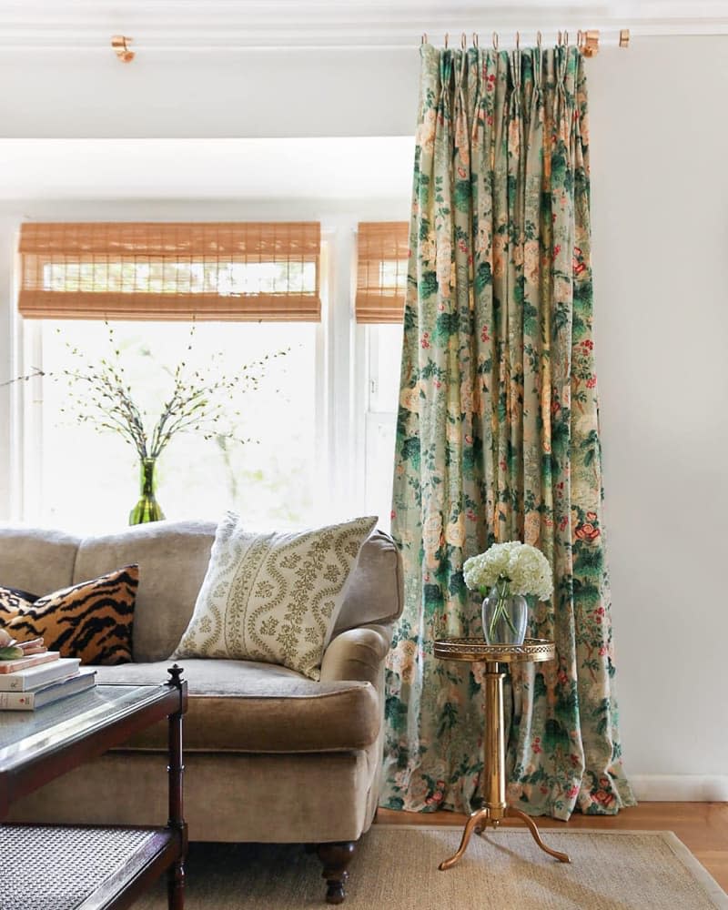 Colorful floral curtain and wood roller shades behind neutral couch and accent table with flowers.