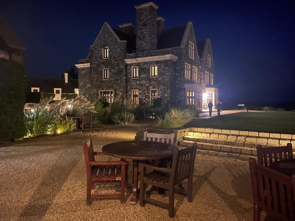 Outdoor patio area with wooden chairs and tables and a home with stone and illuminated windows at nighttime