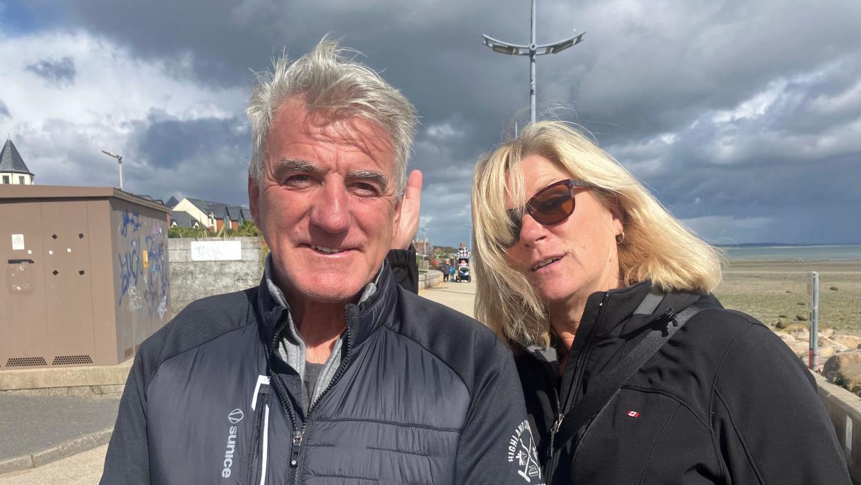 Ted is wearing a grey puff jacket. He is smiling at the camera and has grey hair and a tan.

Julie is mid pose with blonde hair and brown sun glasses. She is wearing a black ski jacket. 

You can see the beach behind and the sea. The sun is shining. 