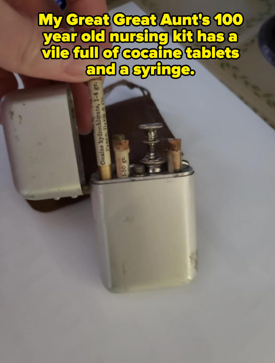 A hand holds an open vintage metal box containing small tubes labeled "Cocaine Hydrochloride," "Heroin," and "Morphine" with a syringe inside