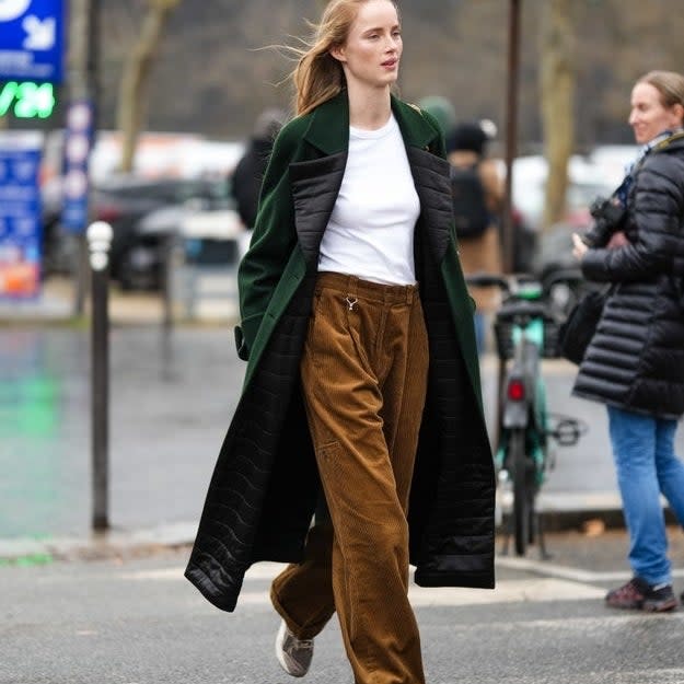 A woman in a long green coat with a black design, white shirt, and brown corduroy pants walks in an urban setting