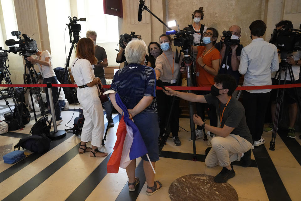 A participant in the attacks trial, holding a French flag, answers reporters as she arrive at the special courtroom Wednesday, Sept. 8, 2021 in Paris. In a secure complex embedded within a 13th-century courthouse, France on Wednesday will begin the trial of 20 men accused in the Islamic State group's 2015 attacks on Paris that left 130 people dead and hundreds injured. (AP Photo/Michel Euler)